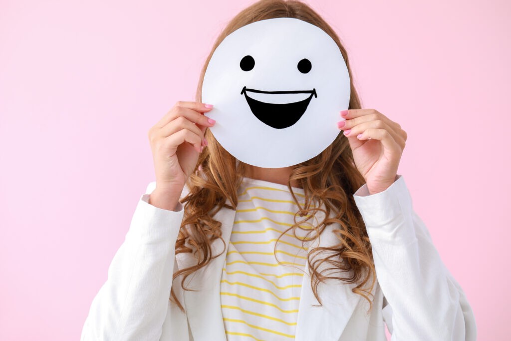 Woman hiding face behind emoticon on pink background