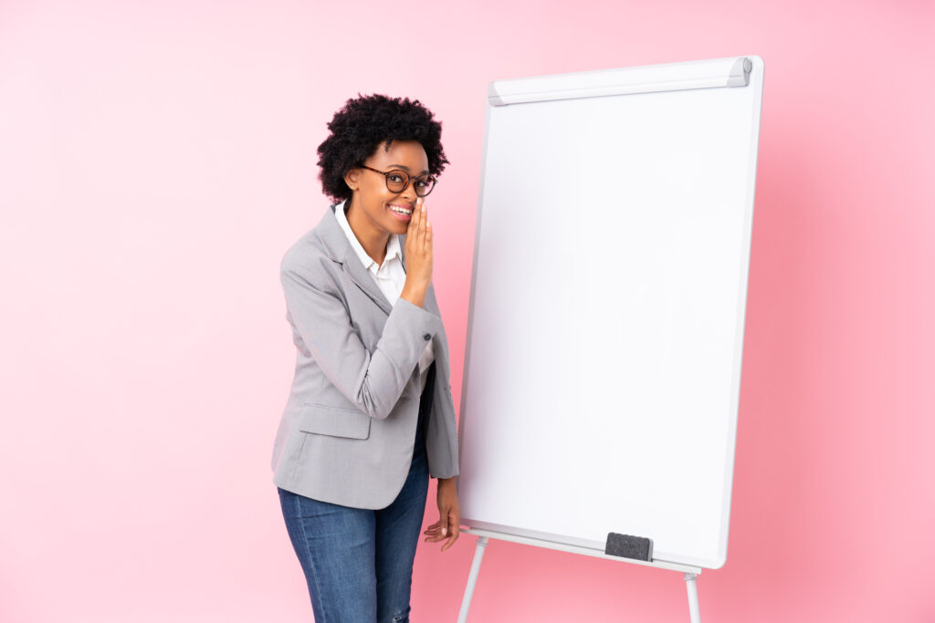 African american business woman giving a presentation on white board over isolated pink background whispering something